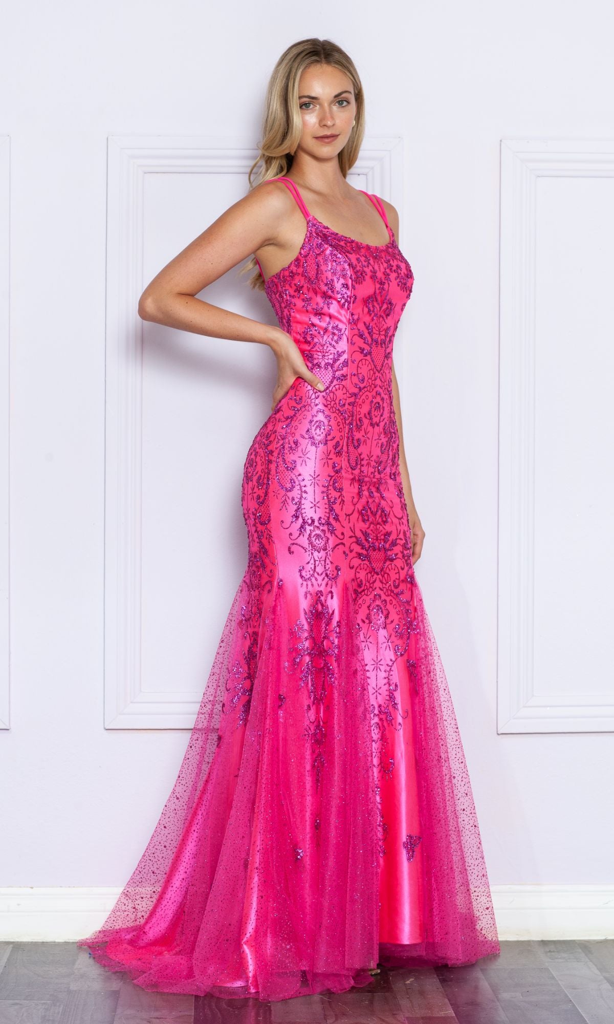 A woman poses sideways against a white paneled wall, wearing the Glitter-Print Long Mermaid Prom Dress 9306—a bright pink, floor-length gown with thin straps and decorative glitter patterns.